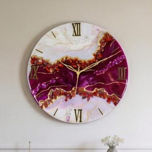 May include: A large circular wall clock featuring a resin art design with swirling shades of deep magenta, white, and cream. The clock face includes gold-colored Roman numerals at the twelve, three, six, and nine o'clock positions and thin gold hands.