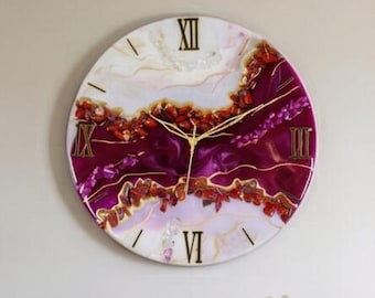 May include: A large circular wall clock featuring a resin art design with swirling shades of deep magenta, white, and cream. The clock face includes gold-colored Roman numerals at the twelve, three, six, and nine o'clock positions and thin gold hands.