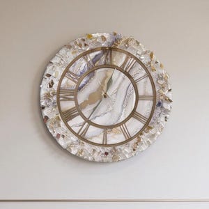 May include: A round wall clock featuring a white and gold marbled face with Roman numerals. The outer frame is decorated with a textured border of clear and iridescent crushed crystals. The clock has gold-coloured hands and a circular gold frame.