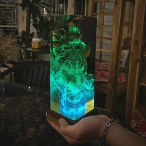 Op de afbeelding: Een rechthoekige harslamp met een houten voet. De lamp heeft een gloeiend groen en blauw ontwerp dat lijkt op rook of gas. De bovenkant van de lamp heeft een donkere, getextureerde uitstraling. De lamp wordt door een persoon vastgehouden.