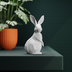 May include: A white rabbit ornament with a geometric design, standing upright with its ears perked. The bunny is on a dark green surface, with an orange pot and green plant in the background.