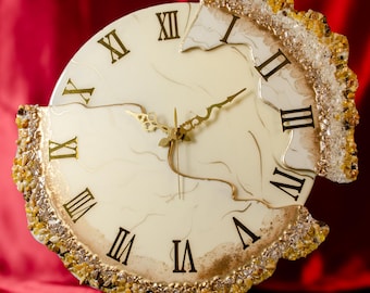 May include: A decorative wall clock with a cream face and gold Roman numerals. The clock's edge is decorated with a textured, amber-coloured material. The clock hands are gold, and the clock is supported by a white stand.