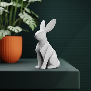 May include: A white geometric rabbit sculpture with long ears, sitting on a dark green surface. The sculpture is next to an orange pot with a green plant. The rabbit has a modern, minimalist design.