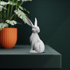 Puede incluir: Una escultura de conejo blanco estilizado, con largas orejas, sentado erguido. La escultura está sobre una superficie verde oscuro, con una maceta naranja y una planta verde en el fondo. El conejo tiene un diseño geométrico moderno.