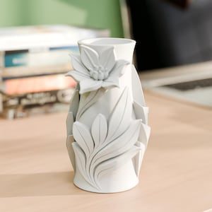 Puede incluir: Un jarrón de cerámica blanco con un diseño floral. El jarrón presenta una flor grande y hojas esculpidas alrededor del cuerpo. El jarrón es cilíndrico con una parte superior acampanada y se asienta sobre una superficie de madera de color claro.