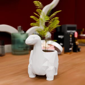 Puede incluir: Una maceta blanca con forma de dinosaurio y diseño geométrico. La maceta contiene una pequeña planta verde con hojas largas y finas. El dinosaurio tiene una cara sonriente y está sobre una superficie de madera.