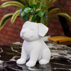 Könnte beinhalten: Ein weißer, hundeförmiger Keramik-Blumentopf mit einem lächelnden Gesicht, der eine kleine grüne Pflanze mit runden Blättern hält. Der Topf steht auf einer dunklen, gemusterten Oberfläche. Die Pflanze verleiht dem Dekor einen Hauch von Natur.