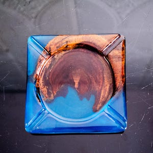 以下が含まれることがあります： 鮮やかな青い樹脂の外装の正方形の灰皿。中央には、濃い茶色の木のような模様と銅色のアクセントが施された円形のデザインがあります。灰皿にはタバコを置くための4つの溝があります。