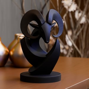Könnte beinhalten: Eine schwarze Skulptur eines Widderkopfes mit großen, gebogenen Hörnern. Die Skulptur hat ein modernes, abstraktes Design mit einem kreisförmigen Sockel und geschwungenen Linien. Der Widderkopf ist zentriert.