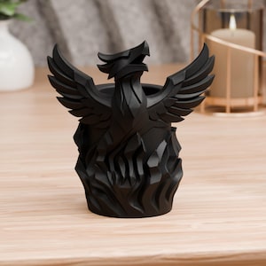 Op de afbeelding: Een zwarte decoratieve kandelaar in de vorm van een feniks met uitgespreide vleugels. De feniks staat op een gestileerde vlammenbasis. De kandelaar is gemaakt van matzwart materiaal en staat op een lichtgekleurd houten oppervlak.