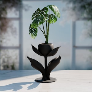 Pode incluir: Um vaso decorativo de metal preto em forma de flor, segurando uma planta Monstera verde. O vaso tem uma base redonda e uma haste com pétalas estilizadas. A planta tem folhas grandes e verdes com buracos distintos.