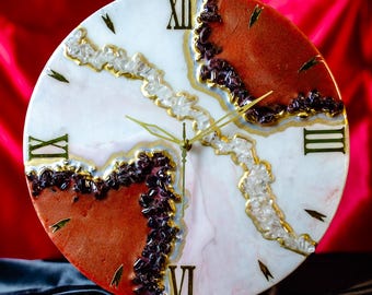 May include: A round decorative wall clock with a white and reddish-brown face. The clock features Roman numerals, gold-colored hands, and decorative accents of clear and dark purple stones. The clock is approximately 12 inches in diameter.