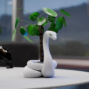 Könnte beinhalten: Ein weißer, schlangenförmiger Keramik-Blumentopf mit einer kleinen grünen Pflanze mit runden Blättern. Die Schlange hat grüne Augen und ist um den Topf gewickelt. Die Pflanze hat einen braunen Stiel.