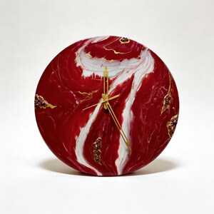 May include: A circular wall clock featuring a glossy, marbled surface with deep red and white swirling patterns. The clock face is accented with small, textured gold and dark red clusters along the edges and includes thin, gold-colored metal hands.