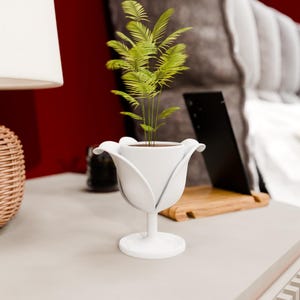 Pode incluir: Um vaso branco em forma de tulipa com uma pequena planta verde dentro. O vaso tem um caule e uma base redonda. A planta tem várias frondes verdes. O vaso está sobre uma superfície cinza claro.