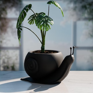 以下が含まれることがあります： 緑の植物が入った黒いカタツムリ型のプランター。 プランターは、スパイラルデザインと2つの触角を備えた丸いボディをしています。 植物は、大きく、緑色で、分割された葉を持っています。 プランターは明るい色の表面に置かれています。