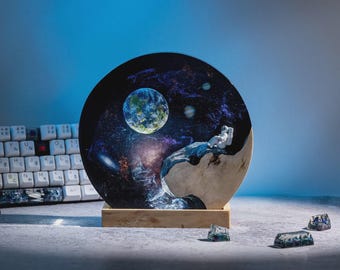 May include: A round, wooden-based art piece depicting a space scene with an astronaut. The artwork features a detailed galaxy, planets, and a seated astronaut. The base is rectangular, and the keyboard keys are visible in the background.