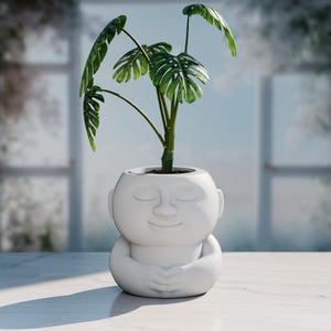 Puede incluir: Una maceta de cerámica blanca con forma de figura meditando, con los brazos cruzados. La maceta contiene una planta verde con hojas grandes y divididas. La maceta está sobre una superficie blanca, con una ventana al fondo.
