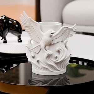 Könnte beinhalten: Eine weiße dekorative Keramikvase mit einer detaillierten Skulptur eines Adlers mit ausgebreiteten Flügeln. Der Adler sitzt über stilisierten Wellen. Die Vase hat eine zylindrische Öffnung und steht auf einer reflektierenden schwarzen Oberfläche.