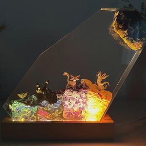 May include: A decorative lamp featuring a clear, angled acrylic panel with a wooden base. Inside, miniature figurines of animals are set amidst colorful, illuminated resin. A large, textured rock is at the top.