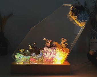 May include: A decorative lamp featuring a clear, angled acrylic panel with a wooden base. Inside, miniature figurines of animals are set amidst colorful, illuminated resin. A large, textured rock is at the top.
