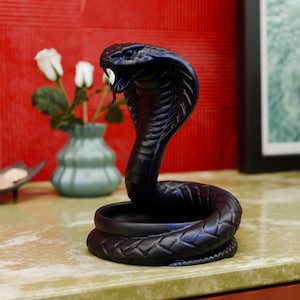Puede incluir: Un objeto decorativo negro con forma de cobra, con la boca abierta y la lengua bífida. La serpiente está enrollada y la cabeza levantada. El objeto está sobre una superficie clara, con un fondo rojo y un jarrón con rosas blancas.