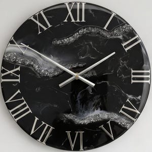 Puede incluir: Un reloj de pared redondo con una superficie de mármol negro y blanco y números romanos plateados. La esfera incluye detalles de purpurina plateada texturizada en un acabado de resina, con manecillas plateadas finas y rectas.