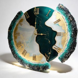 May include: A circular wall clock featuring a deep emerald green and white marbled resin face with gold Roman numerals. The clock has a textured, jagged outer edge encrusted with dark green and black crystalline accents and thin gold clock hands.