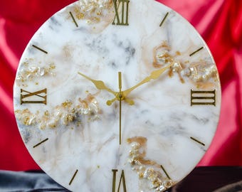 May include: Circular wall clock featuring a white marble-effect face, gold Roman numerals, and gold hands. The clock face has gold embellishments and is set against a red backdrop. The clock is supported by a white stand.