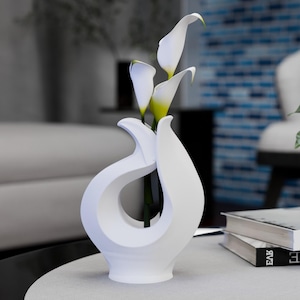 May include: A white, abstract ceramic vase with a unique, flowing design, holding several white calla lilies with yellow centers. The vase sits on a light-coloured surface, with a blurred background.