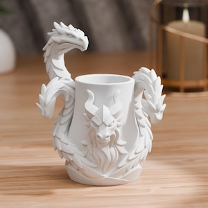 Pode incluir: Um recipiente branco de cerâmica com tema de dragão. O recipiente apresenta três cabeças de dragão, com duas formando alças e uma centralizada na frente. Os dragões têm escamas e chifres detalhados. O recipiente é cilíndrico com um acabamento liso e fosco.