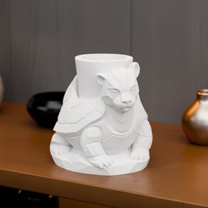 May include: A white decorative figurine featuring a stylized bear-turtle hybrid design. The sculpture has a cylindrical opening and is made of a solid, matte white material. The bear-turtle is sitting on a geometric base.