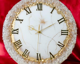 May include: A circular decorative clock with a white face, gold Roman numerals, and gold hands. The clock has a gold rim and is surrounded by clear crystals. The background is a deep red.