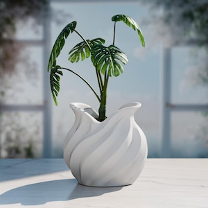 Puede incluir: Un jarrón de cerámica blanco, con remolinos, que contiene una planta de Monstera con hojas grandes, verdes y divididas. El jarrón tiene un diseño escultórico único. La planta está colocada frente a una ventana con un fondo borroso.