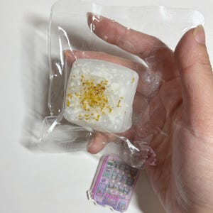 May include: A square, white confection with yellow flecks, sealed in clear plastic. A small, colorful sticker of a vending machine is visible below the confection. The items are held in a hand against a white background.