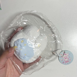 Puede incluir: Un juguete blando blanco en forma de nube con un centro azul claro que presenta un diseño de luna y estrellas. El juguete está empaquetado en una bolsa de plástico transparente e incluye una etiqueta redonda con un personaje de dibujos animados.