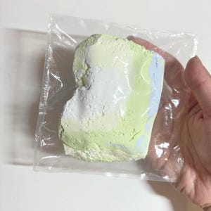 May include: A colorful, textured art piece in a clear plastic bag. The piece features a mix of white, light green, and light blue colors. The art piece is held in a person's hand.