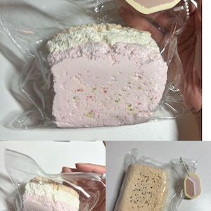 May include: A decorative cake slice in a clear plastic bag, featuring layers of pink, white, and tan. The cake has a textured surface and is decorated with tiny, colorful sprinkles. A small tag with a cake slice design is attached.