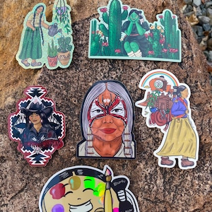 May include: A selection of vibrant stickers showcasing Indigenous women and related imagery. Designs include a woman watering plants, a woman with cacti, a woman in a cowboy hat, a woman with face paint, a woman with a cart, and a smiling artist with a paintbrush.