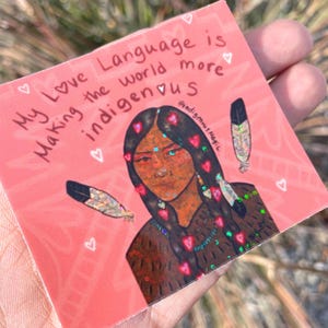 May include: A square sticker with a pink background featuring an illustration of a woman with long black hair and a brown top. The text "My Love Language is Making the world more indigenous" is written on the sticker, along with two feathers and hearts.