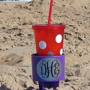 Pode incluir: Um porta-copos roxo ancorado na areia com um monograma branco "D.H." em um círculo prateado. Um copo vermelho com bolinhas brancas e um canudo vermelho está colocado em cima do porta-copos.