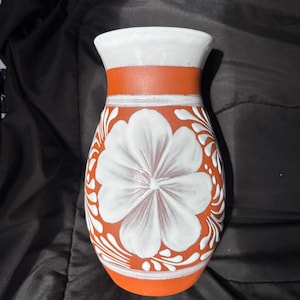 Könnte beinhalten: Eine handgefertigte Keramikvase mit einem weißen Blumendesign auf einem terrakottafarbenen Körper. Die Vase hat einen weißen Rand und ein weißes Band um den Hals. Das Blumendesign ist eine große, stilisierte Blume.