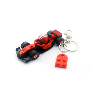 May include: Red toy race car keychain with black wheels and a driver wearing a white-striped helmet. The keychain includes a red brick-shaped charm and a silver metal clasp.