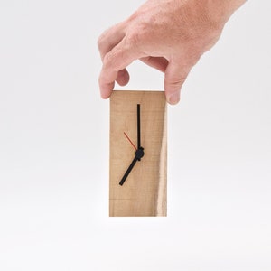Peut inclure: Une horloge en bois avec des aiguilles noires et une aiguille des secondes rouge. L'horloge est tenue par une main.