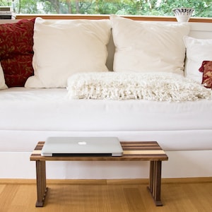 May include: A white couch with red and white pillows and a white throw blanket. A laptop sits on a wooden table in front of the couch.
