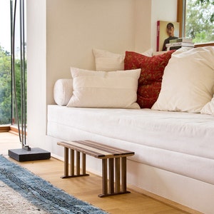May include: A white couch with pillows and a wooden bench in front of it. The couch is against a white wall and there is a window behind it. The bench is made of wood with a natural finish and has a simple design.
