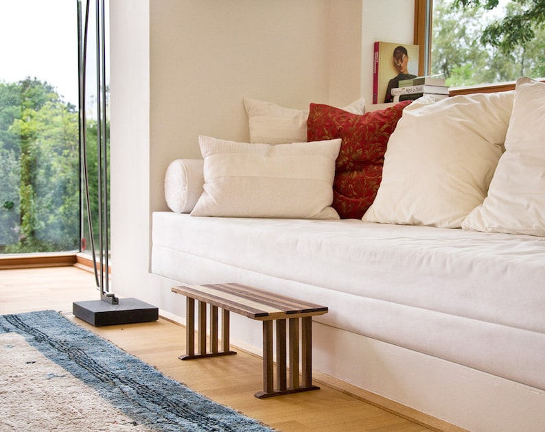 May include: A white couch with pillows and a wooden bench in front of it. The couch is against a white wall and there is a window behind it. The bench is made of wood with a natural finish and has a simple design.
