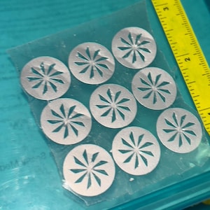 以下が含まれることがあります： 星型のデザインが施された、銀色の円形金属片9個のコレクション。各ピースには、中心から放射状に切り抜かれた形状があります。これらの部品は透明なビニール袋に入っており、黄色の定規の隣にあります。