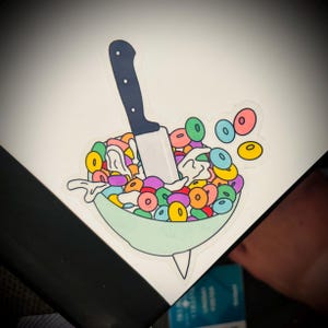 May include: A sticker featuring a bowl of colorful cereal with a knife stabbed through it. The cereal is a mix of red, orange, yellow, green, blue, and purple. The bowl is light green, and the knife has a black handle.