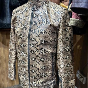 以下が含まれることがあります： 茶色とベージュのヘビ柄レザー ジャケット。黒いジッパーと襟が付いています。 ジャケットはスタンドカラーとスリムフィットです。 袖口にはジッパーが付いています。 ジャケットはマネキンに展示されています。